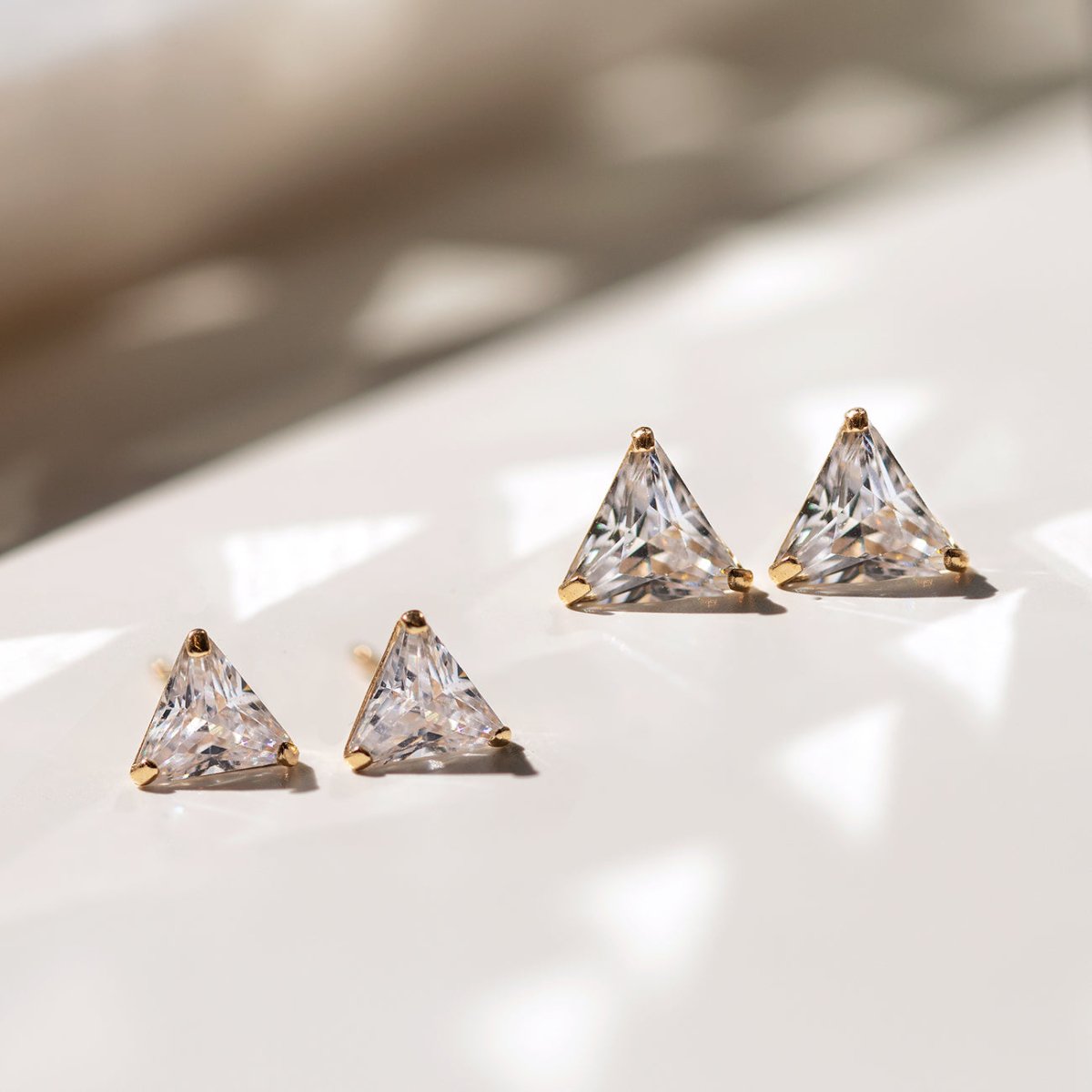 14K gold triangle white zirconia studs in two sizes, on a white surface in sunlight