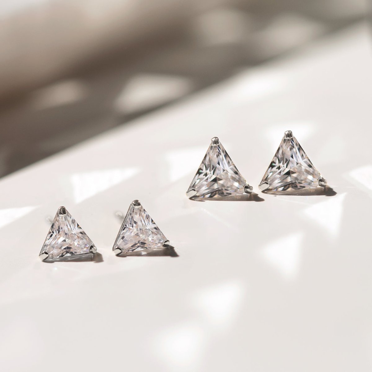 14K white gold triangle white zirconia studs in two sizes, on a white surface in sunlight