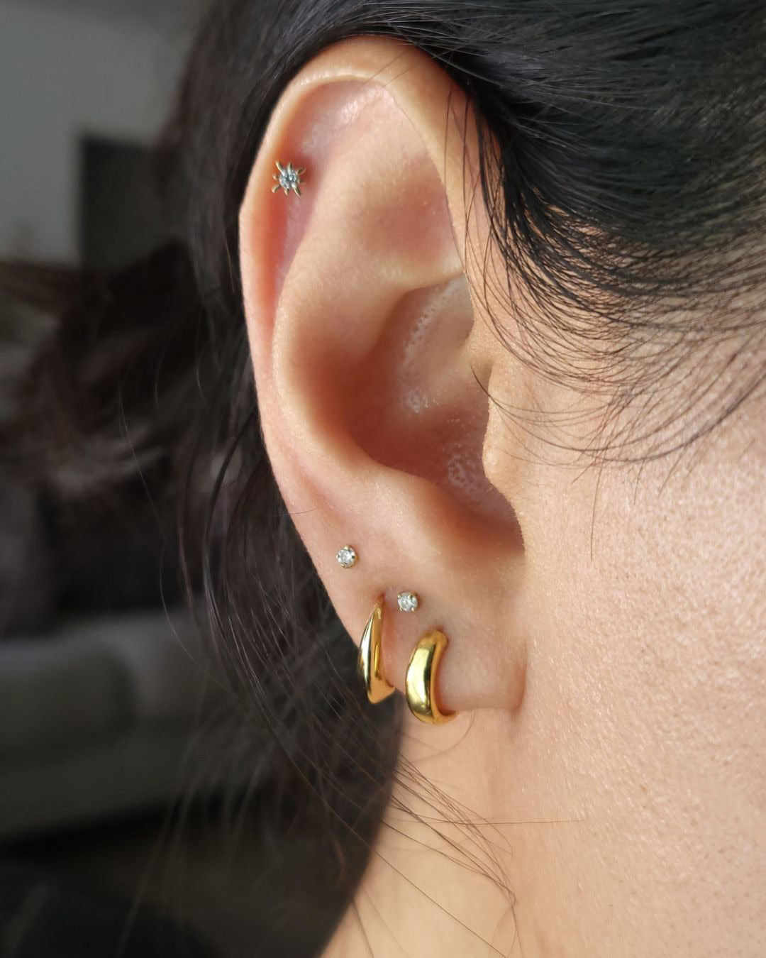 Close-up of an ear wearing a gold starburst crystal stud in the helix, two crystal studs and two gold huggie hoops