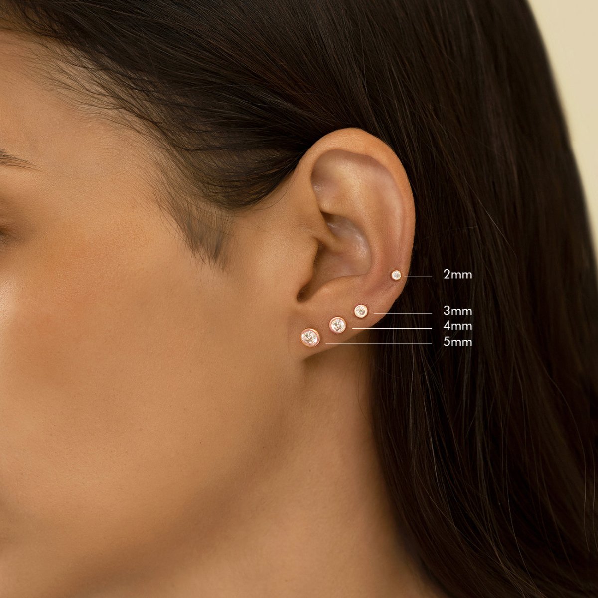 14K rose gold bezel zirconia studs on a model in a size guide marked 2mm, 3mm, 4mm and 5mm