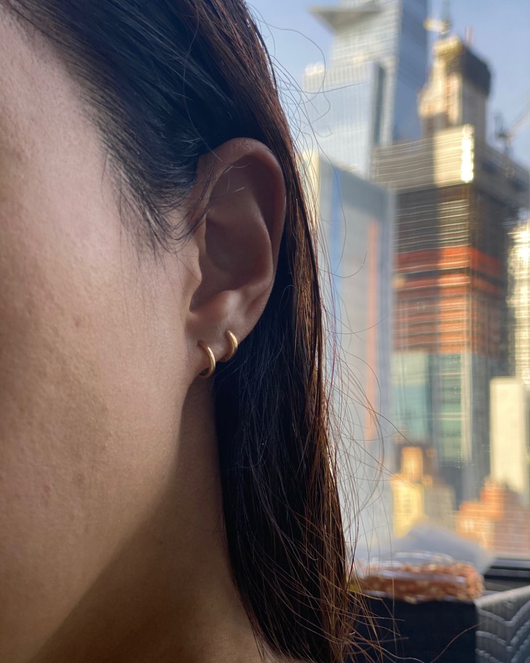 Two small gold huggie hoops worn in stacked lobe piercings, with a blurred city skyline behind