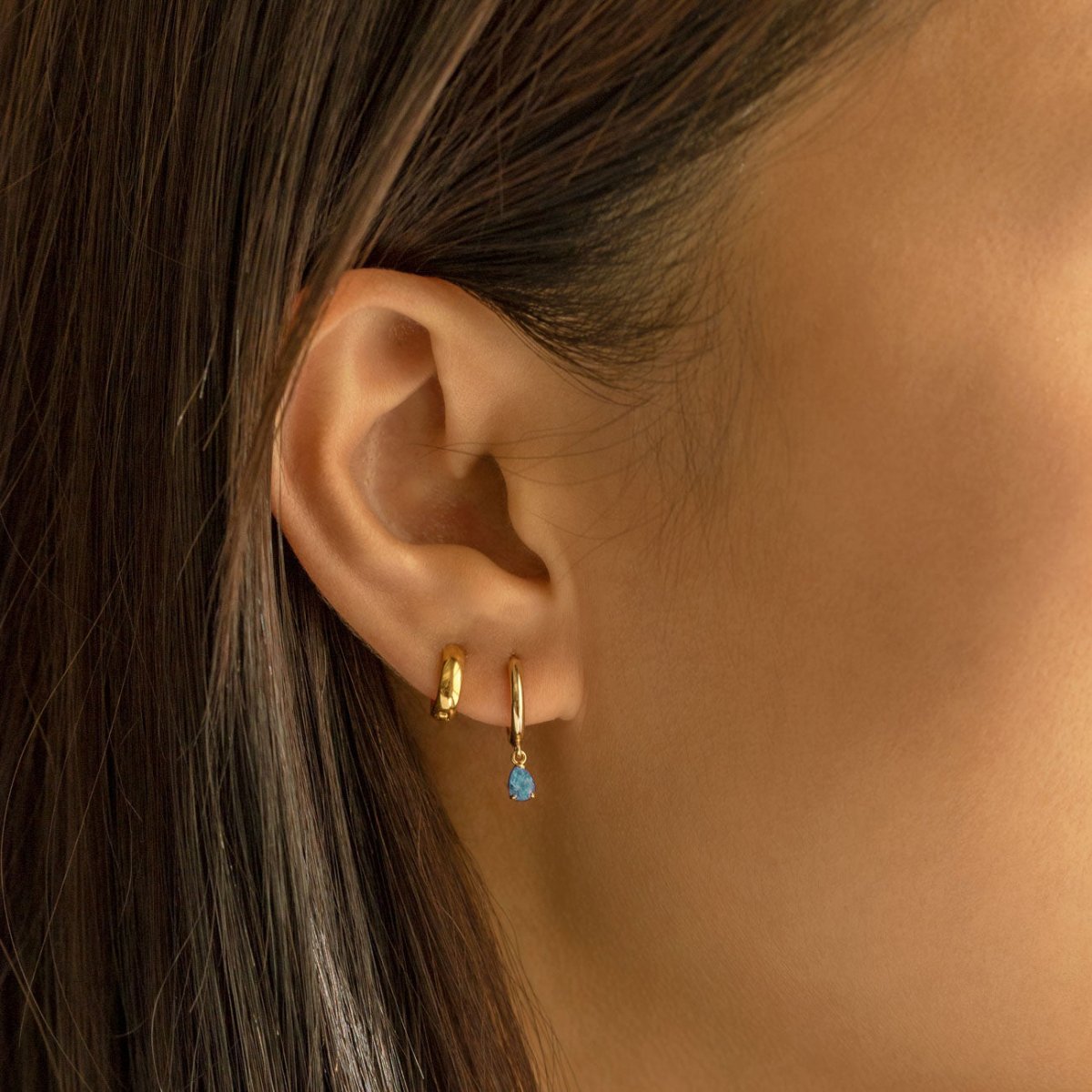 14K gold huggie hoop with a blue zircon drop, shown on a model's lobe