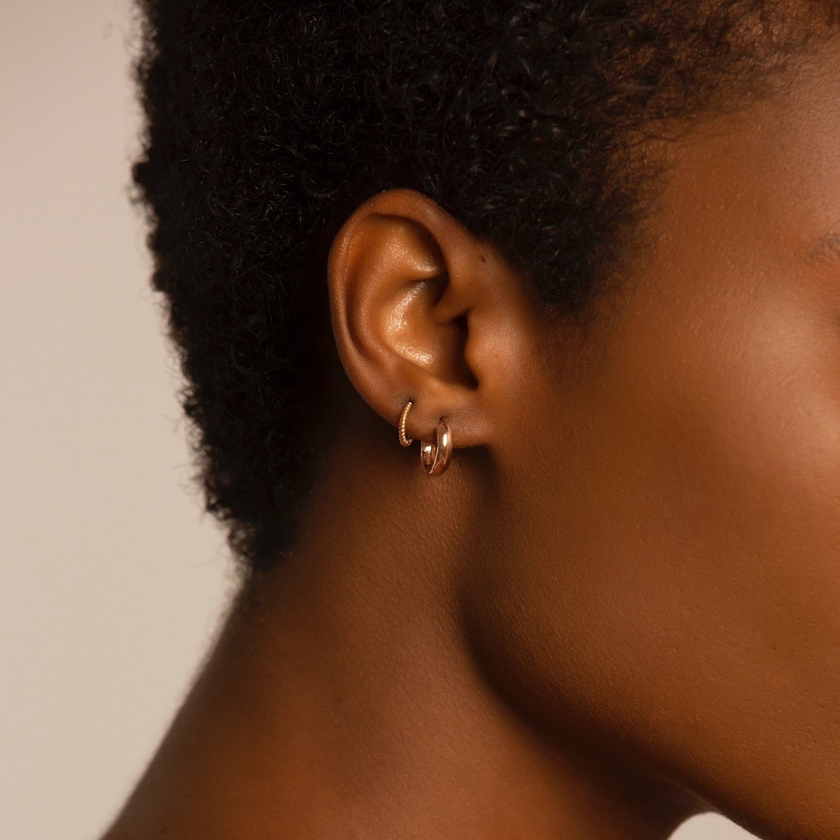 14K rose gold mini huggie hoops shown on a model's lobe, viewed from behind the ear