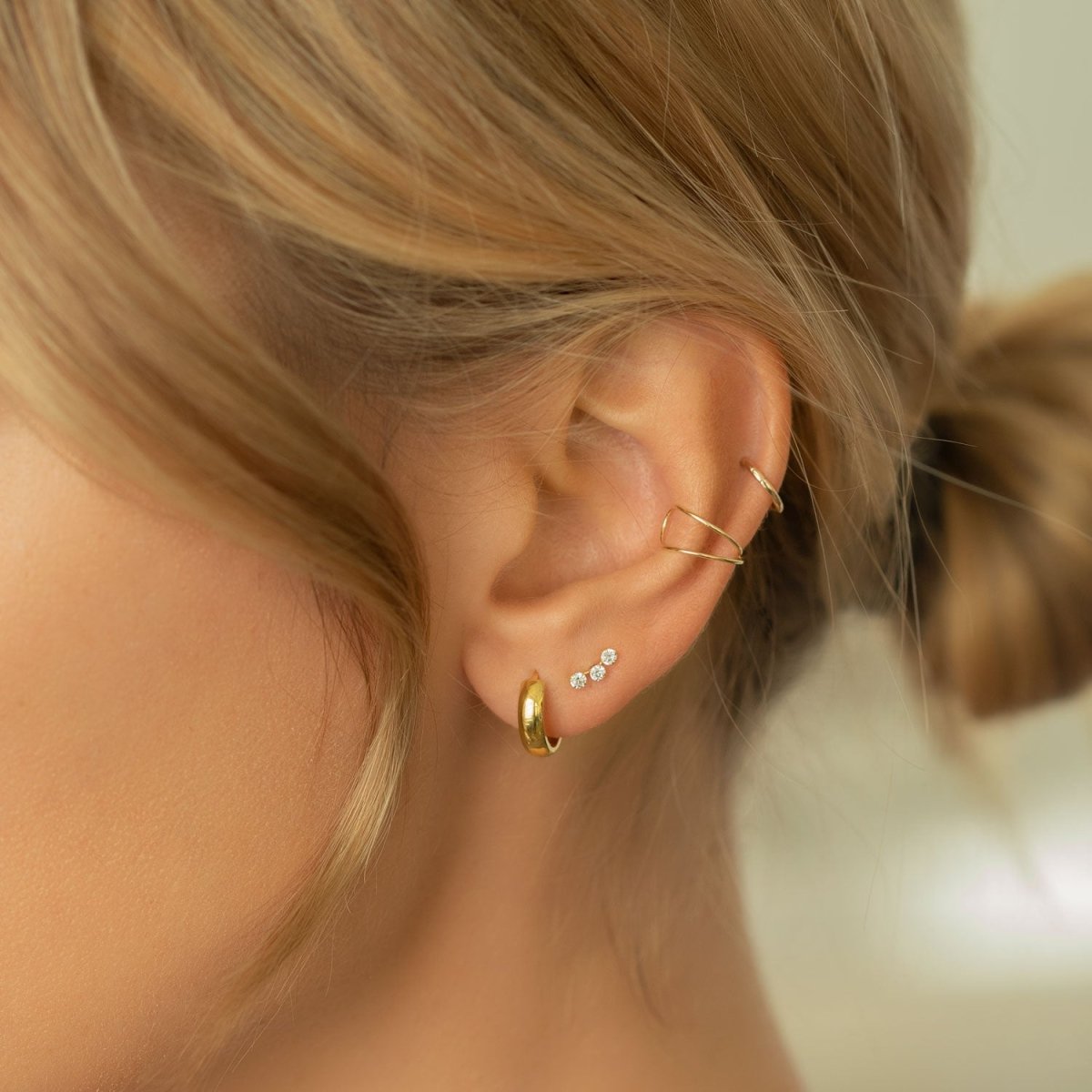 Close-up of woman's ear featuring 14k gold boyfriend mini hoops, 14k curve pave bar stud, 14k duo ear cuff, and 14k thin huggie hoop