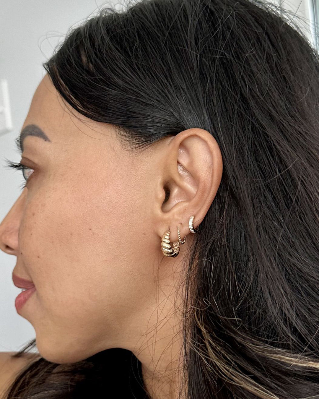 Model's ear worn with a chunky ribbed gold hoop in the lobe and two smaller twisted hoops above