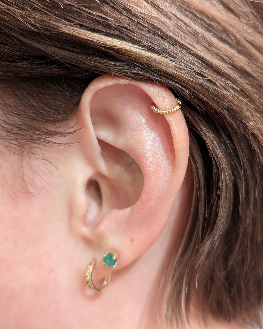 Close-up of an ear wearing a beaded gold helix hoop, a green stone lobe stud and a small gold hoop