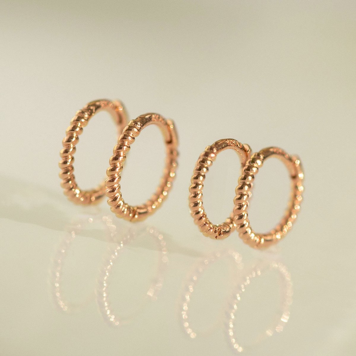 Two pairs of rose gold vermeil twisted rope huggie hoops, on a reflective surface