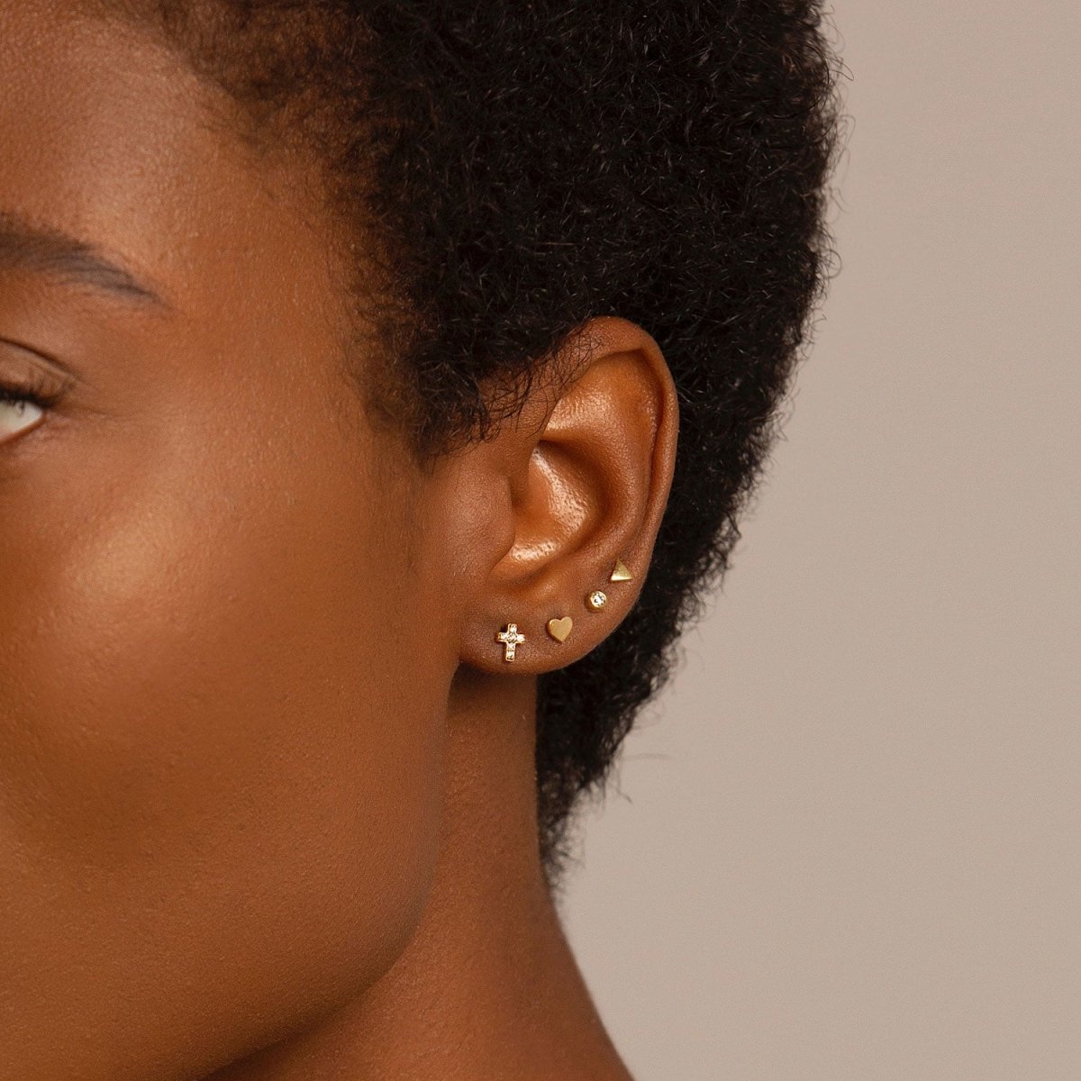 14K gold diamond bezel stud styled in an ear stack with cross and heart studs, on a model's lobe