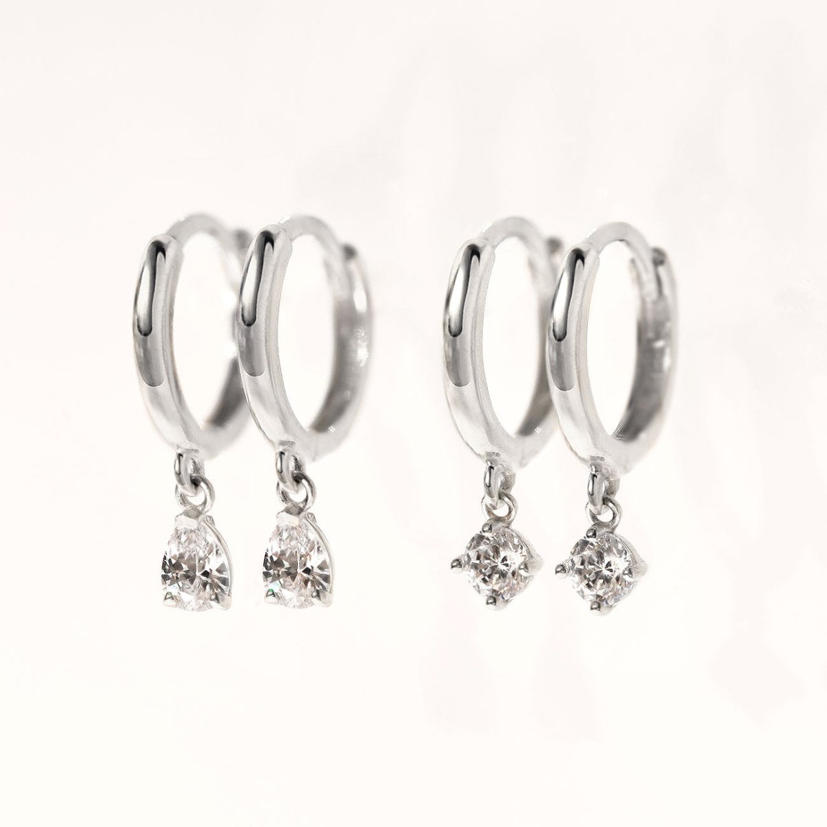 14K white gold pear-drop moissanite huggies beside round moissanite huggies, on a cream background