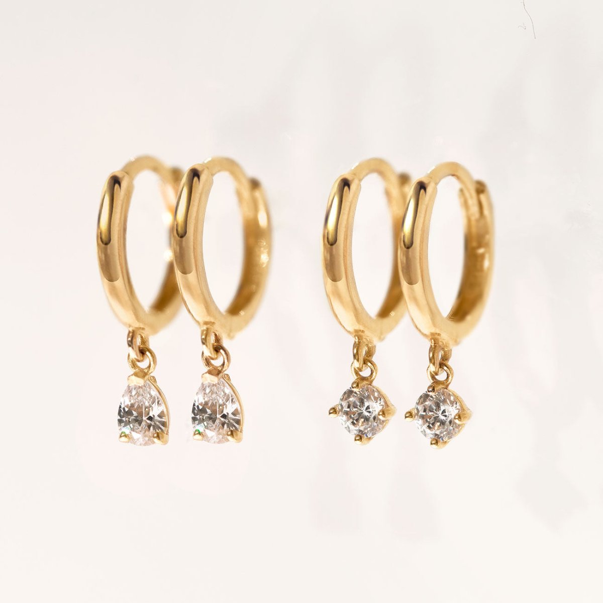 14K gold pear-drop moissanite huggies beside round moissanite huggies, on a cream background