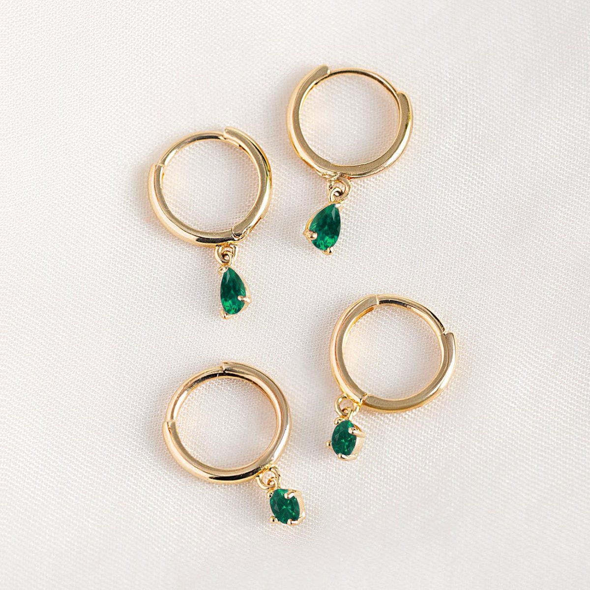 14K gold huggie hoops with pear-shaped green emerald drops, shown loose on a cream background