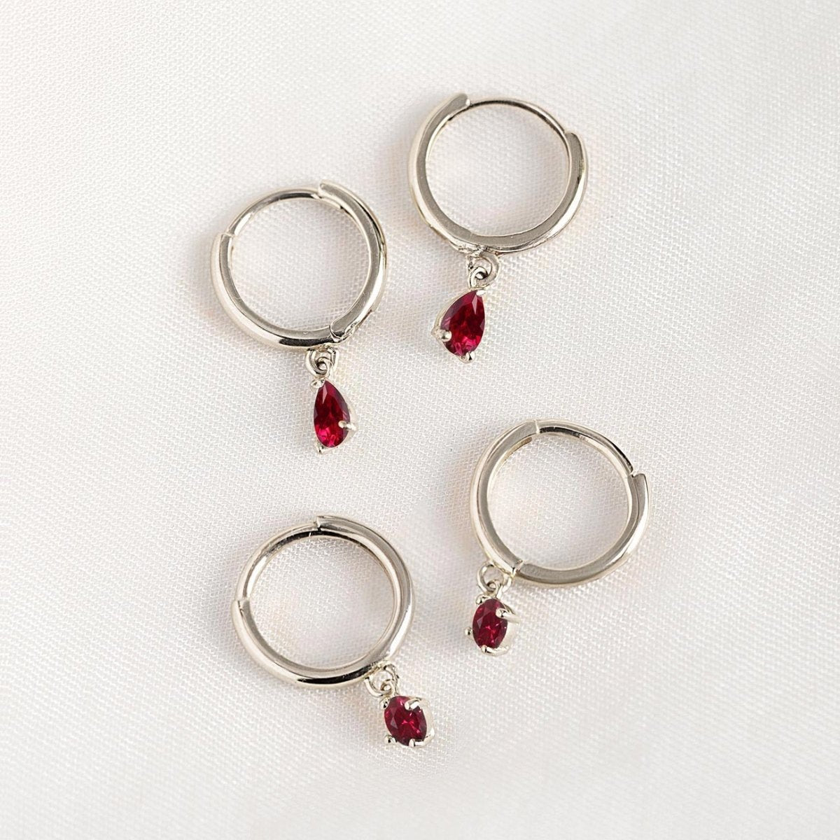 14K white gold huggie hoops with pear-shaped red ruby drops, shown loose on a cream background