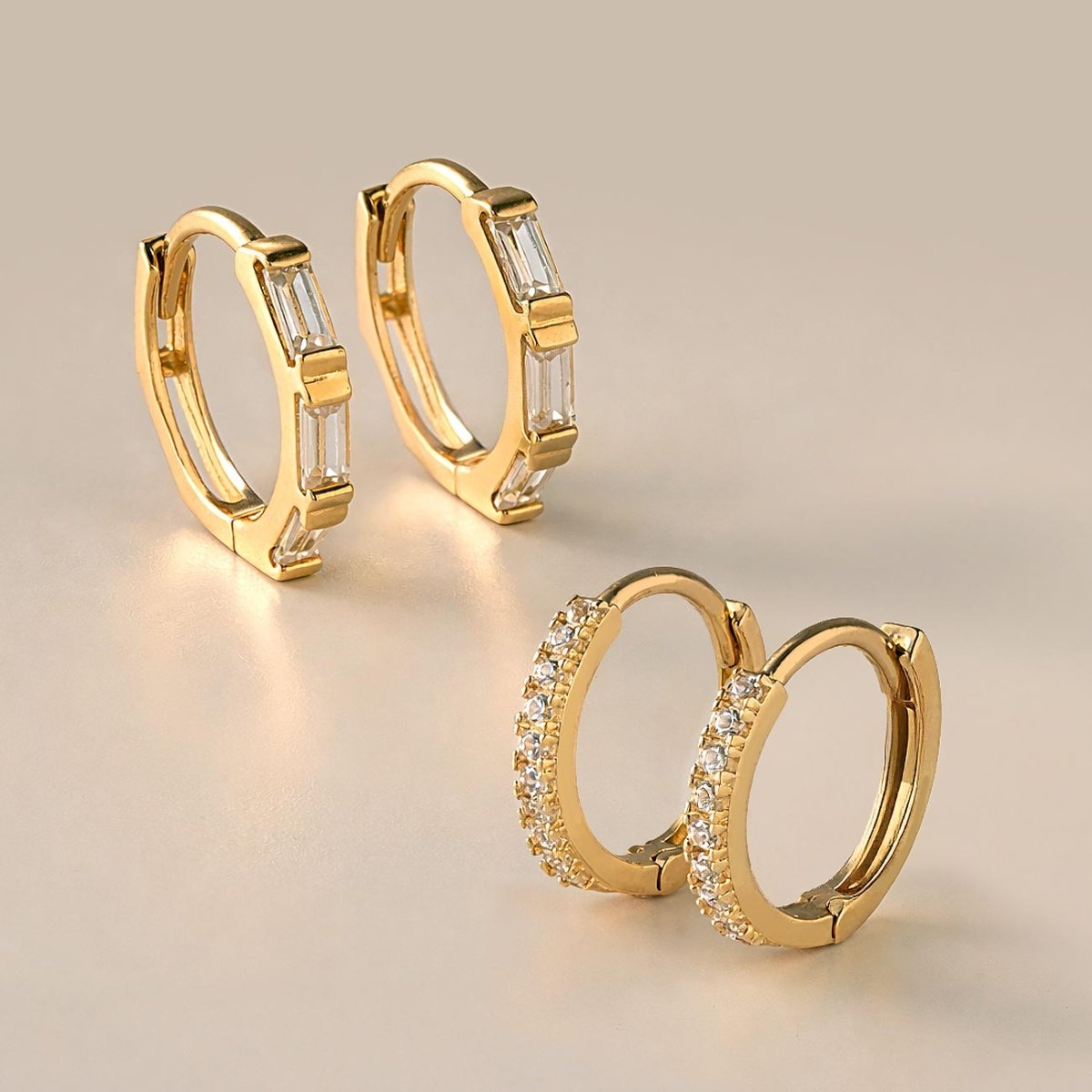 14K gold baguette huggie hoops with clear stones, beside pave huggies on a cream background