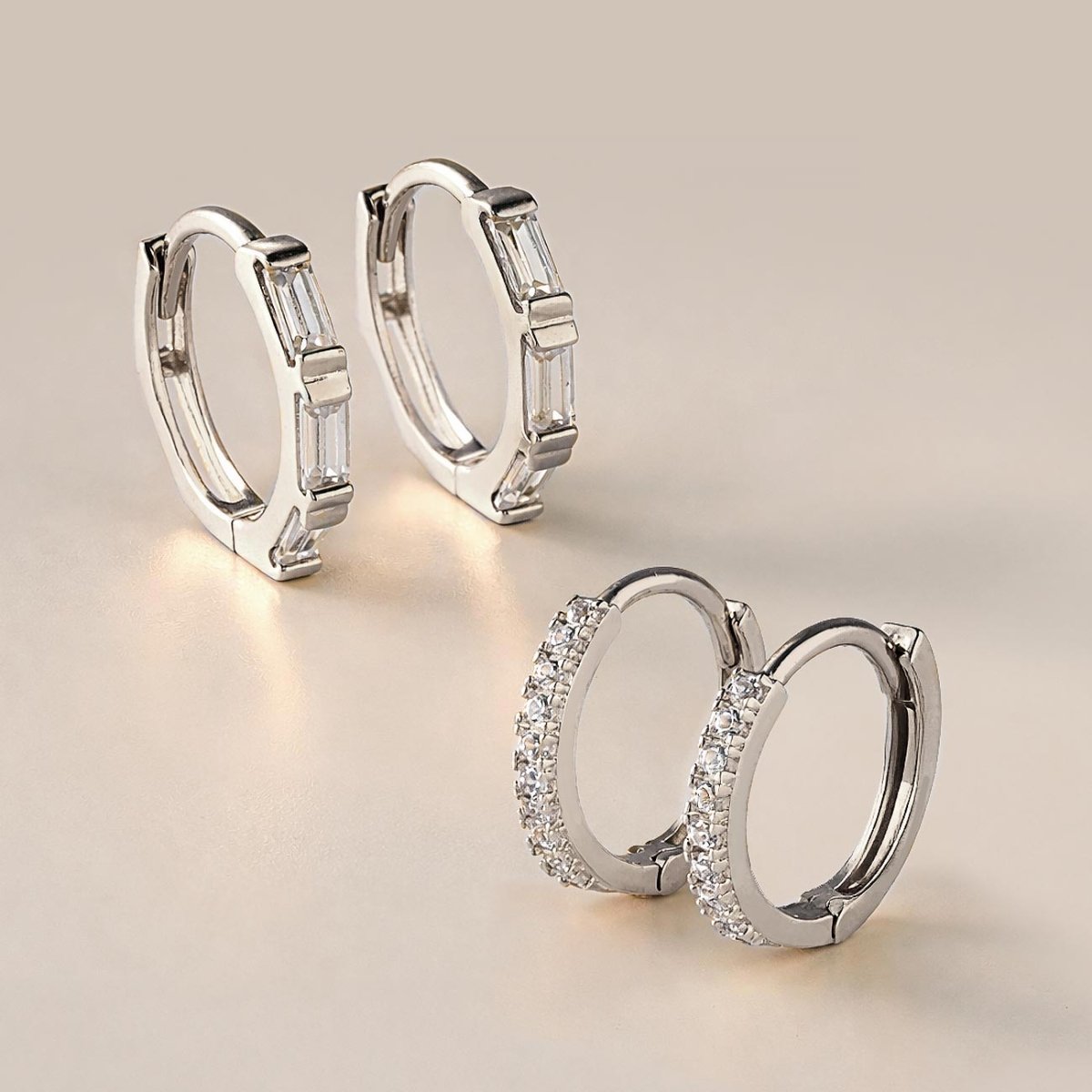 14K white gold baguette huggie hoops with clear stones, beside pave huggies on a cream background
