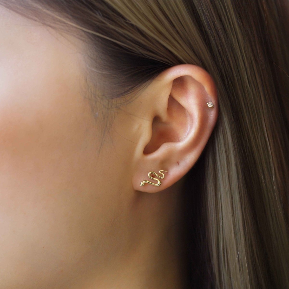 14K gold serpent stud earring shown on a model's lobe, with a tiny clear stud on the helix