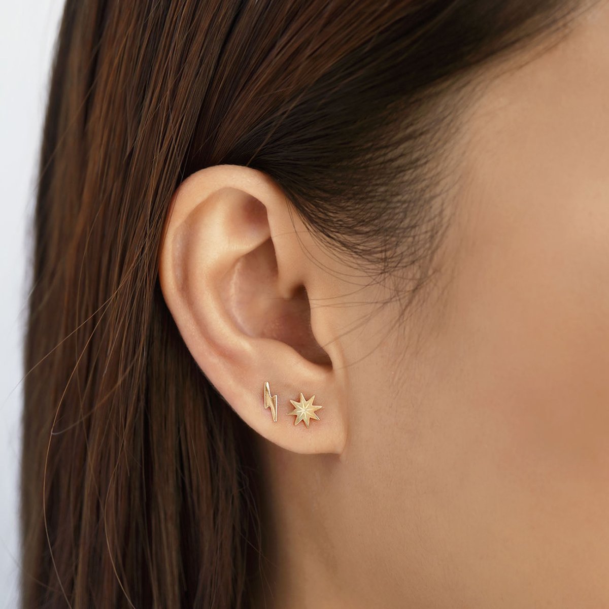 14K gold tiny lightning bolt stud worn on a model's lobe beside a separate starburst stud