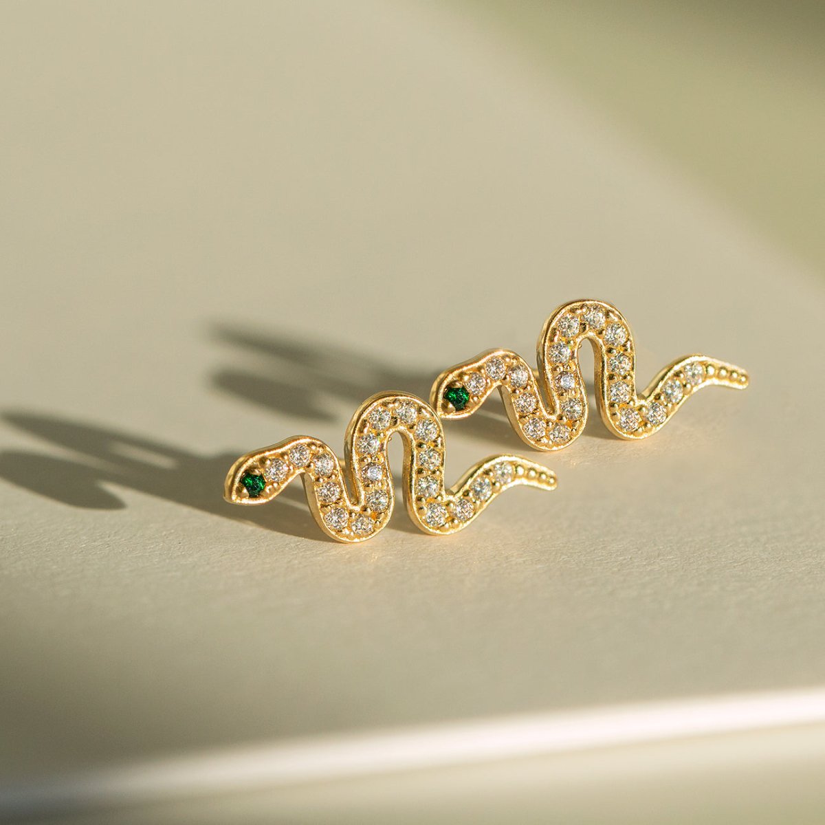 14K gold pavé snake studs with clear zirconia and green stone eyes, close-up detail on a cream background