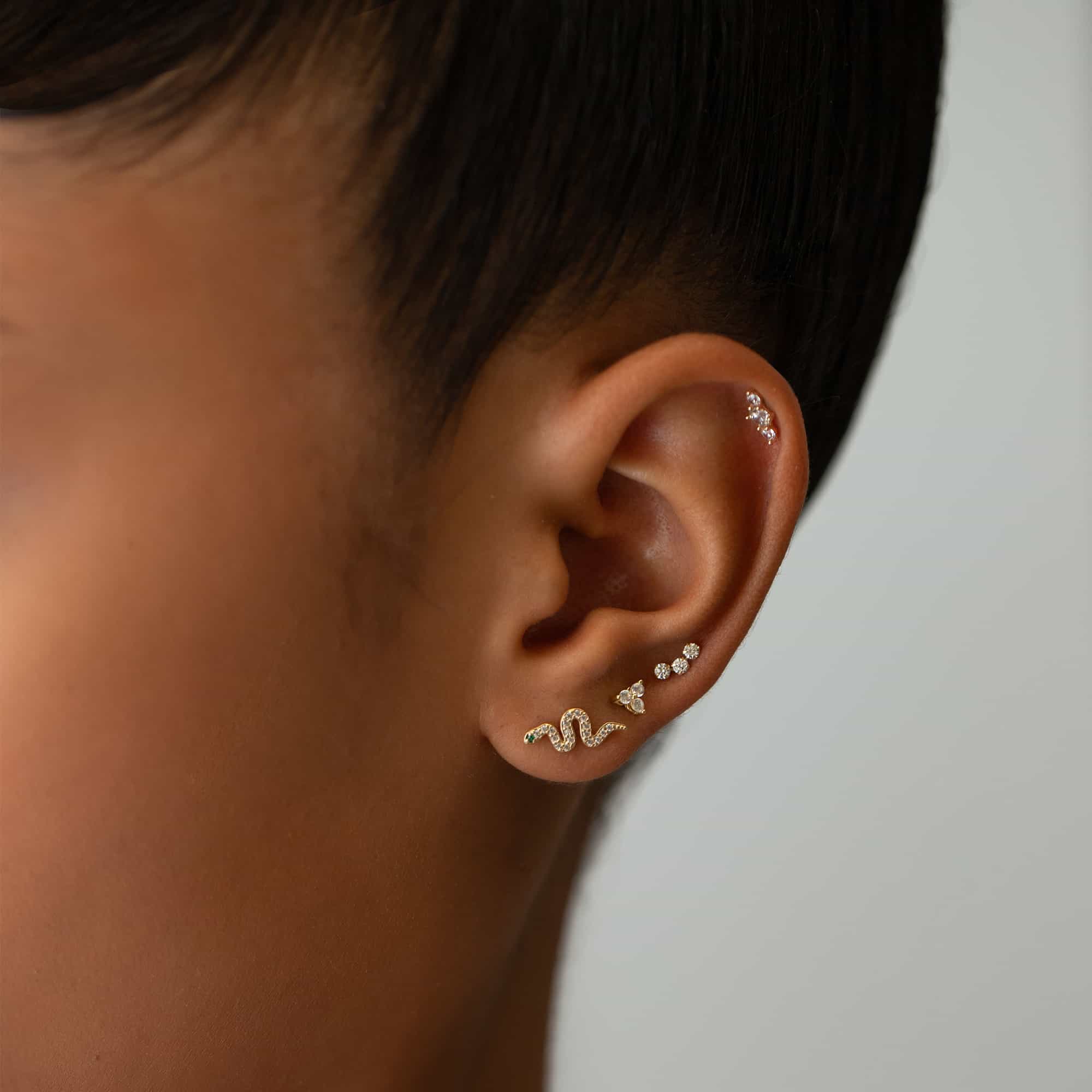 Close-up of woman's ear featuring 14k gold snake pave studs, 14k gold tiny clover pave stud, 14k gold curve pave bar stud, and 14k gold trio bar pave stud