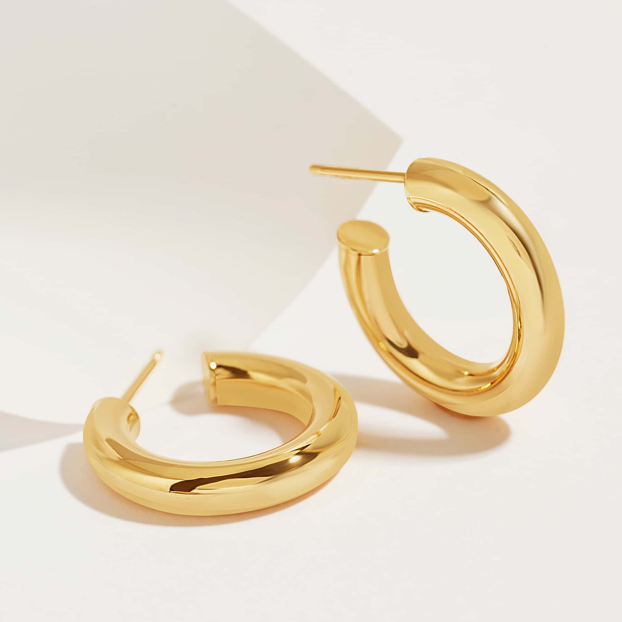 14K gold thick chunky hoop earrings shown at an angle on a cream surface