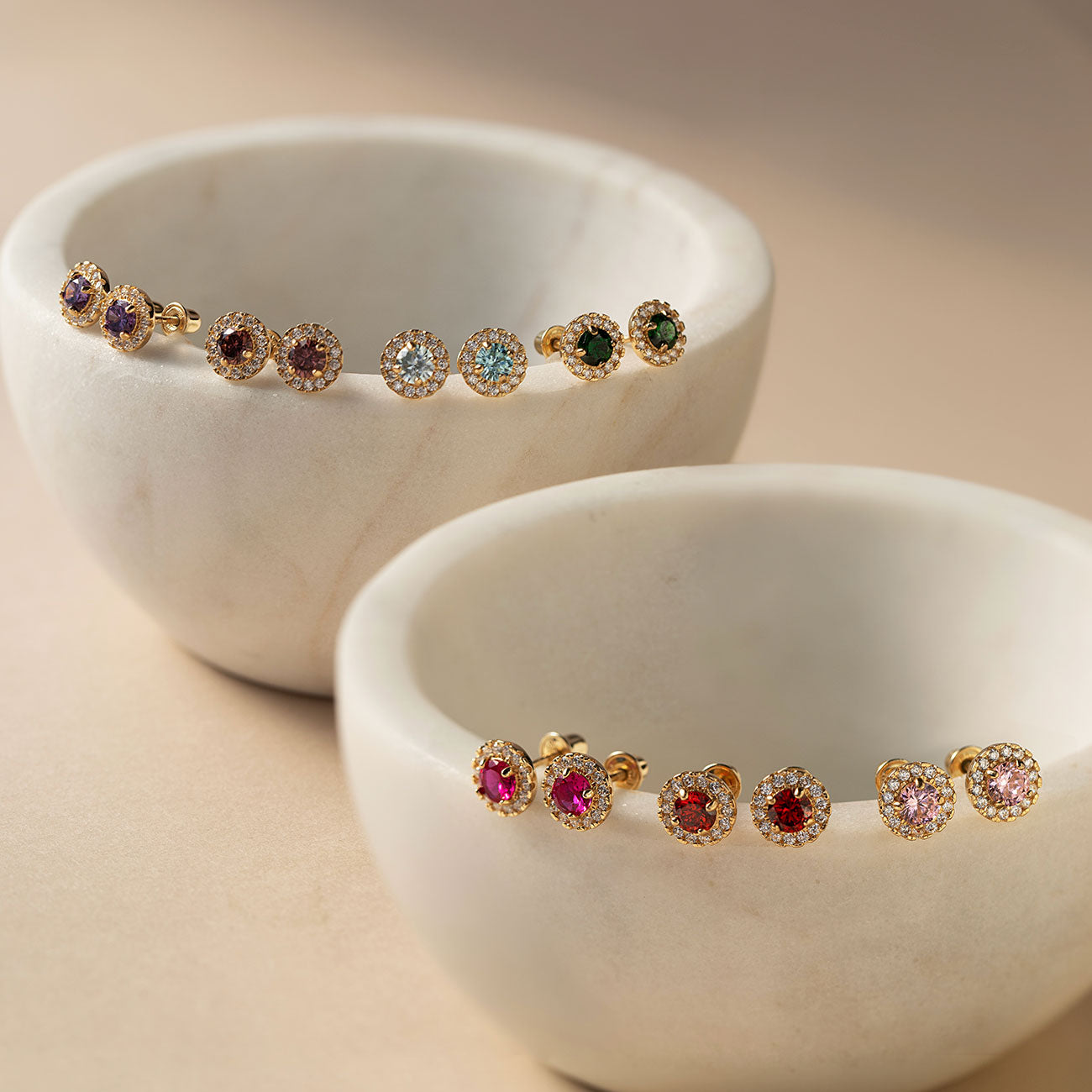 Colorful solitaire halo stud earrings form AMYO featuring assorted birthstone gemstones displayed on white bowls against a beige background.