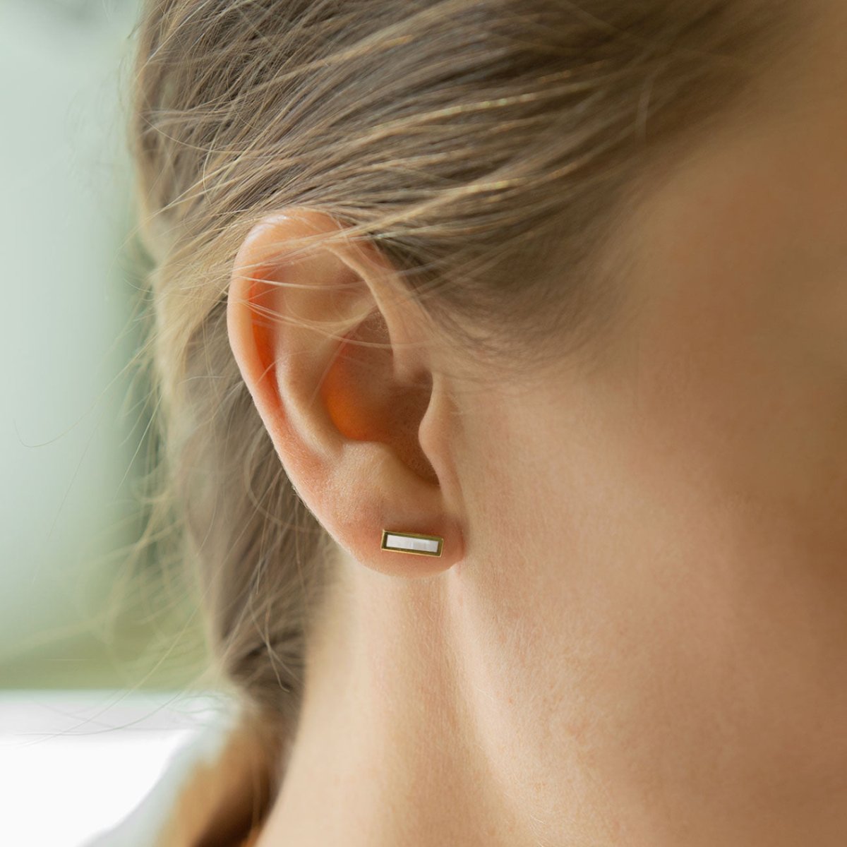 14K gold mother of pearl bar stud with a pale iridescent stone, shown on a model's lobe