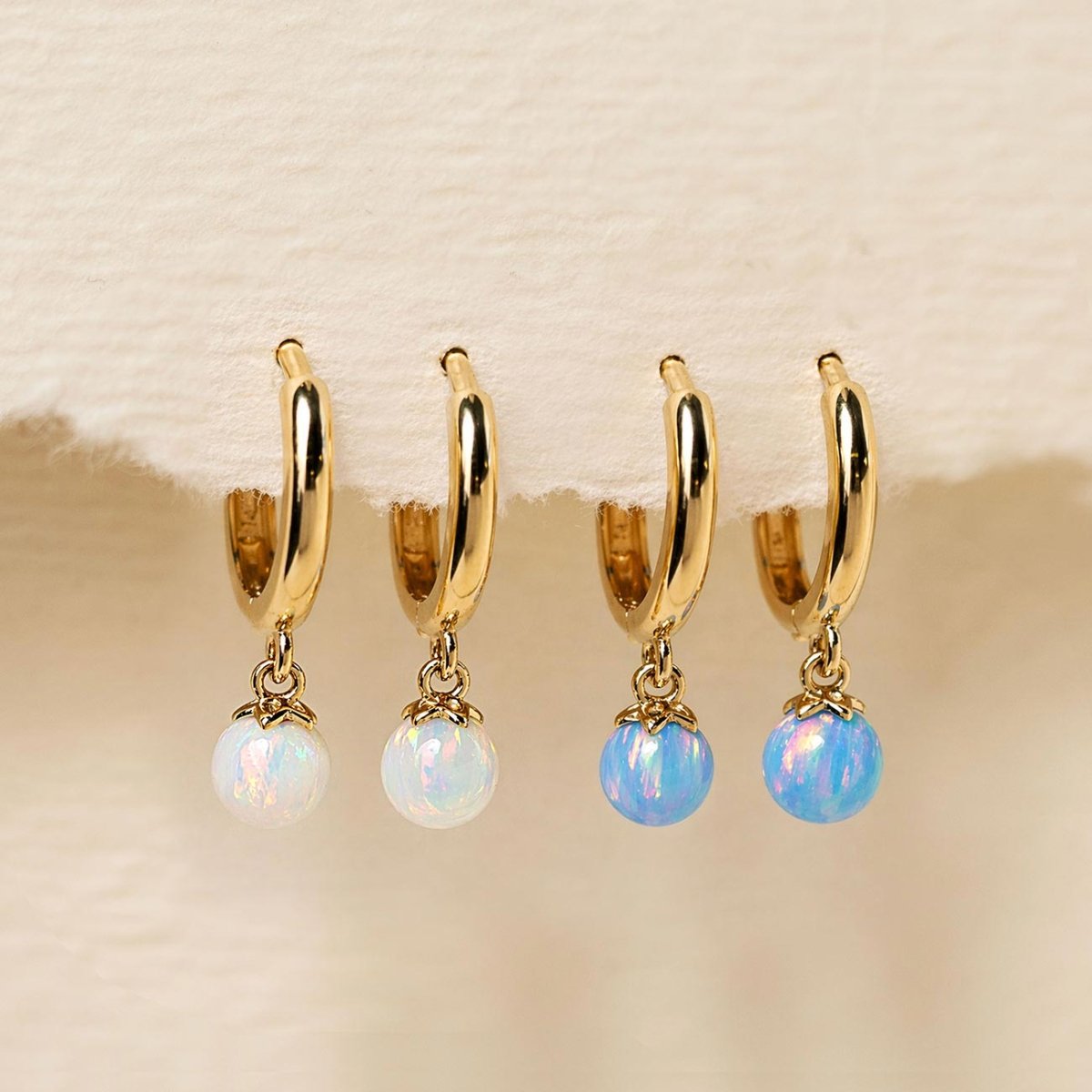 14K gold opal huggie hoops shown in white and light blue opal beads, on a cream background
