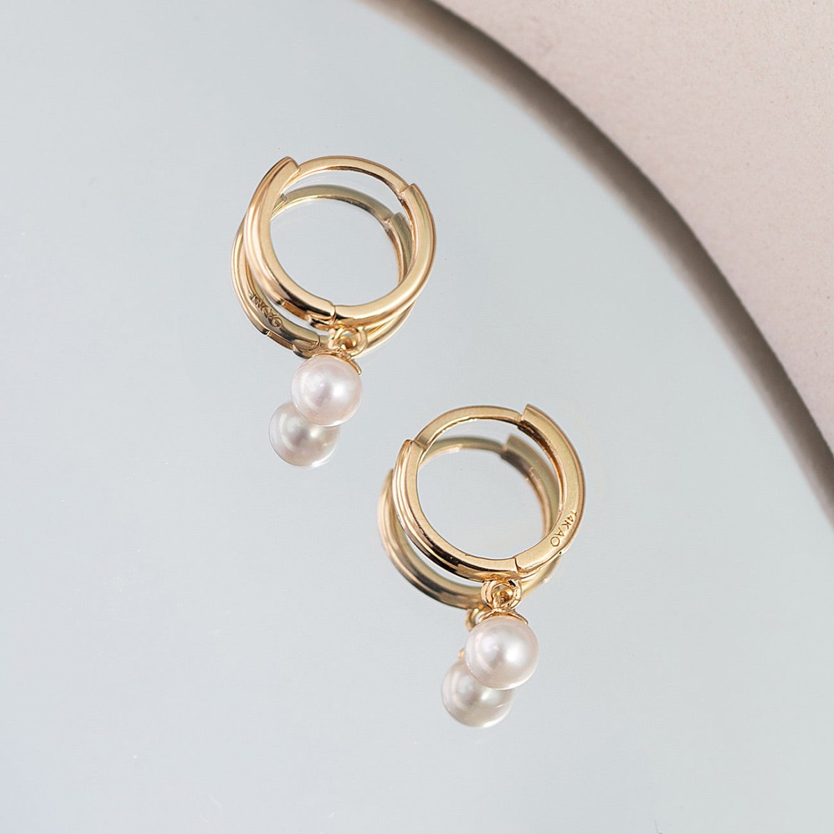 14K gold huggie hoop earrings with two pearl drops, on a pale gray surface