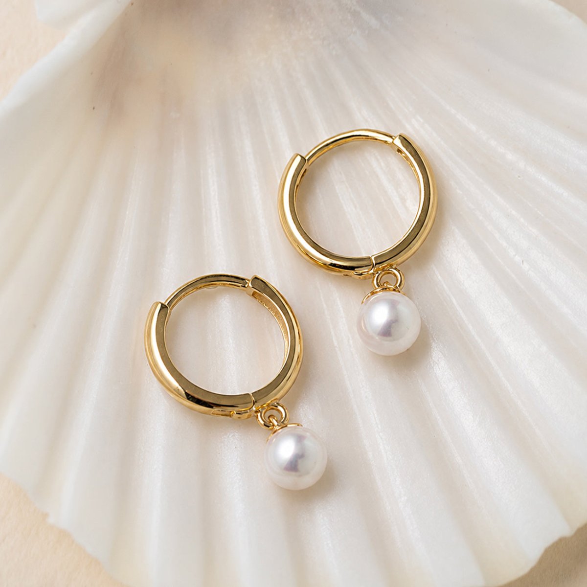 Pair of 14K gold huggie hoops with dangling pearls on a white shell background.