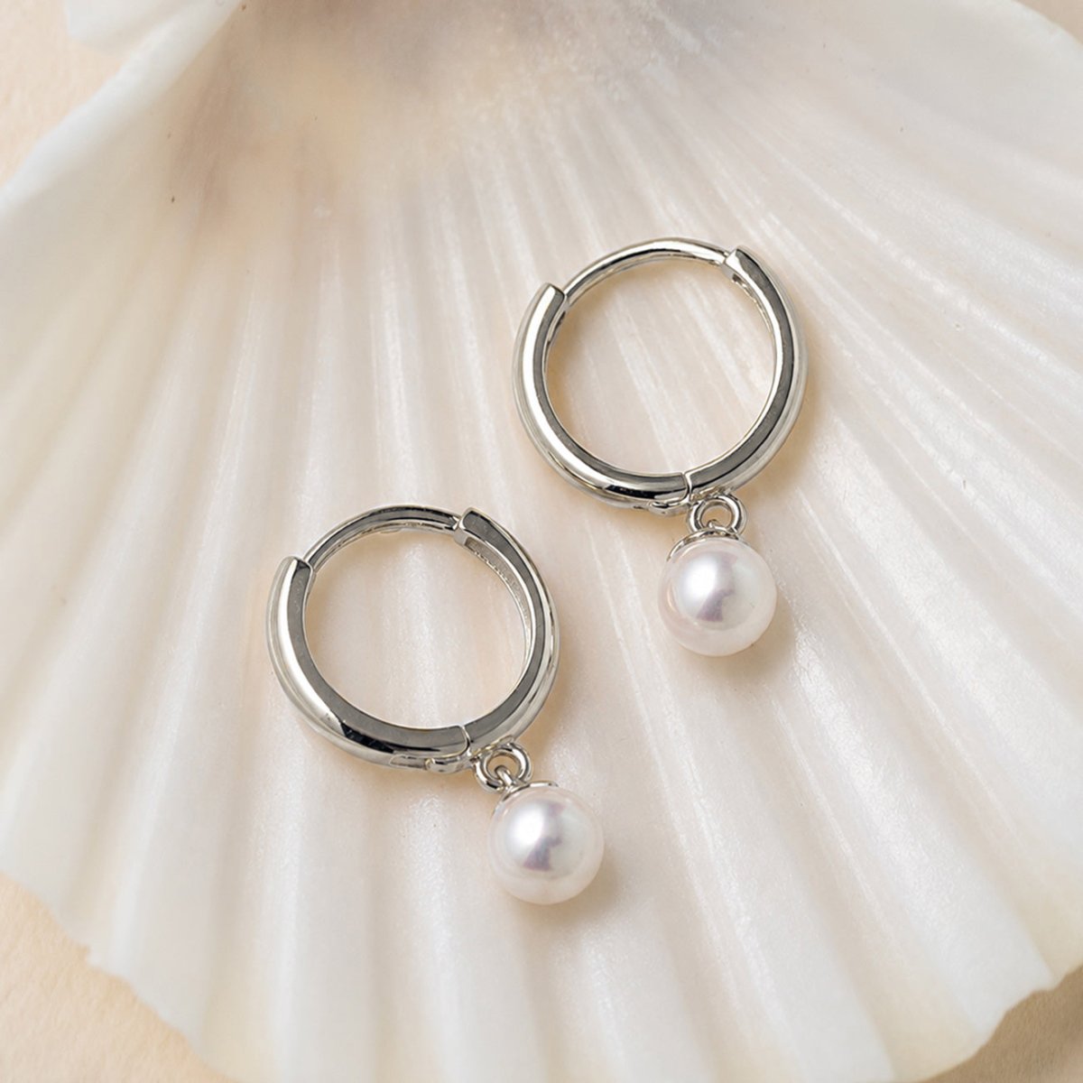 Pair of 14K white gold tiny huggie hoops with dangling pearls on a white shell background.