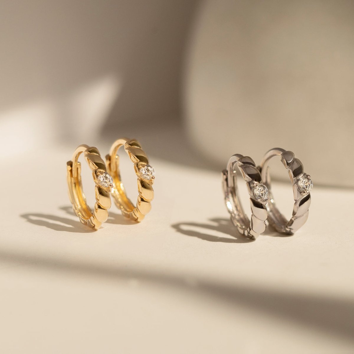 14K white gold rope twist huggie hoops with clear stones beside gold pairs, on a cream background