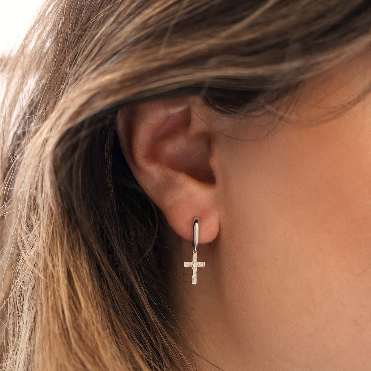 14K white gold huggie with a dangling crucifix cross charm, shown close-up on a model's lobe