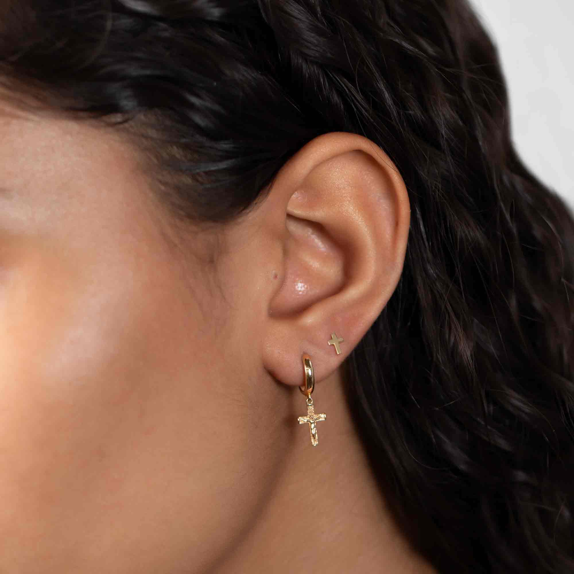 14K gold cross stud shown on a model's ear stacked above a hoop with a crucifix drop