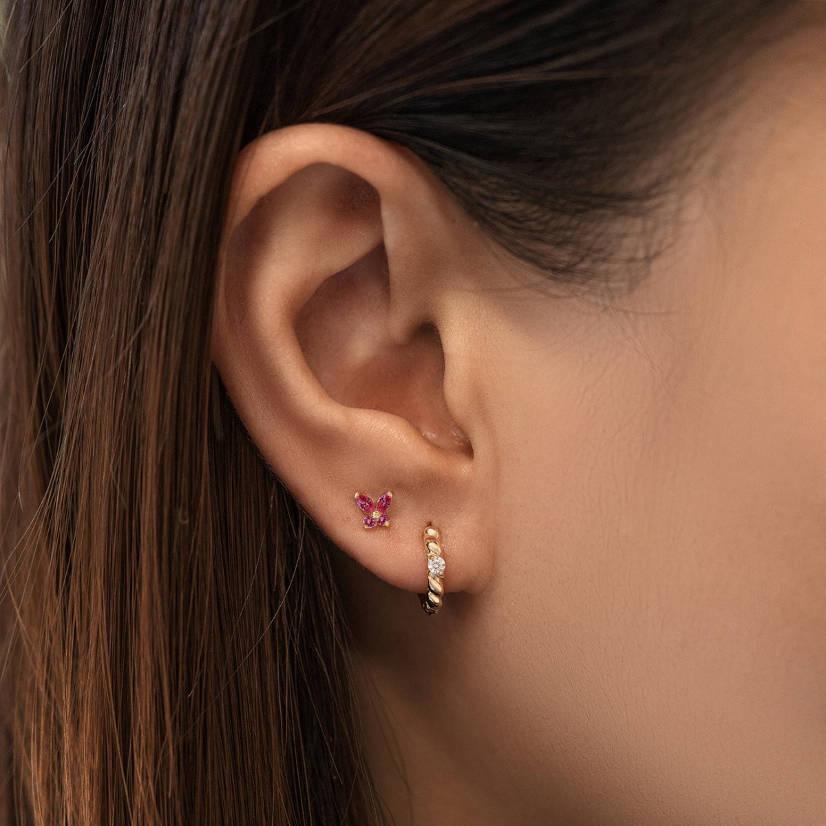 14K gold ruby butterfly stud with red stones beside a twisted huggie hoop, on a model's lobe