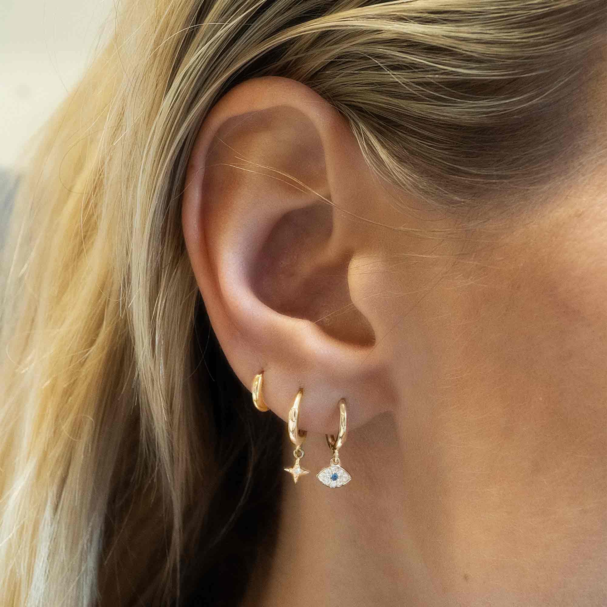 14K gold evil eye huggie hoop with pave clear and blue stone charm, shown stacked on a model's lobe