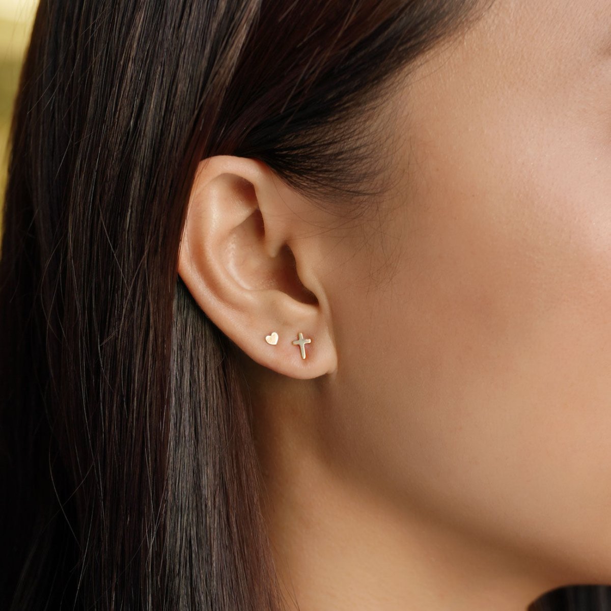Gold-filled cross stud and a small heart stud shown on a model's lobe, close-up