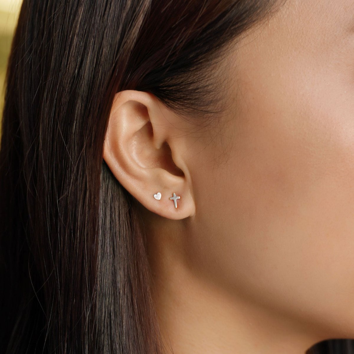 14K white gold cross stud beside a tiny heart stud, shown on a model's lobe