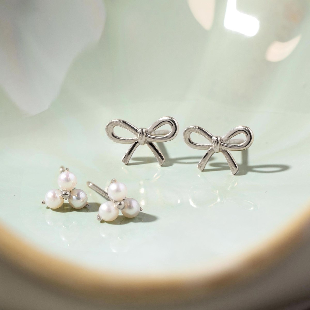 14K white gold trinity pearl studs shown with bow studs in a green dish