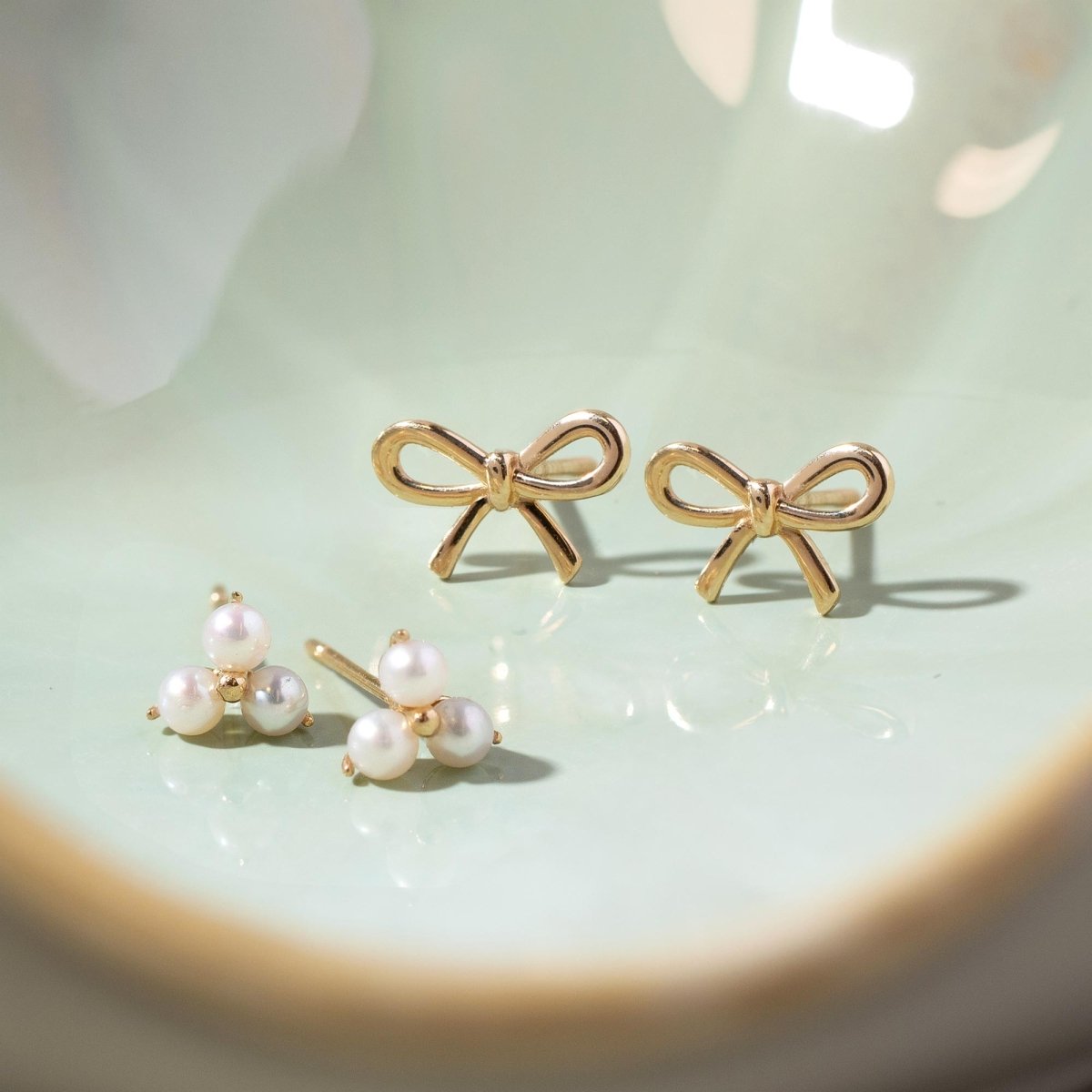 Close up of earrings featuring 14K Gold Bow Studs and pearl clover studs 