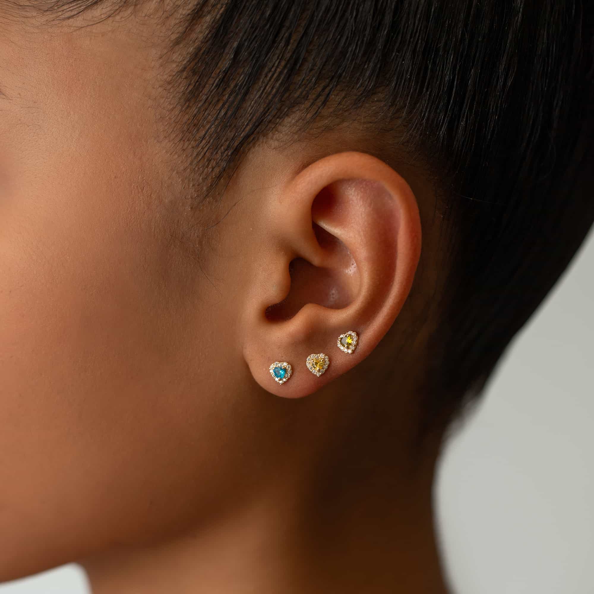 14K gold peridot heart halo stud shown on a model's lobe in a stack of heart studs