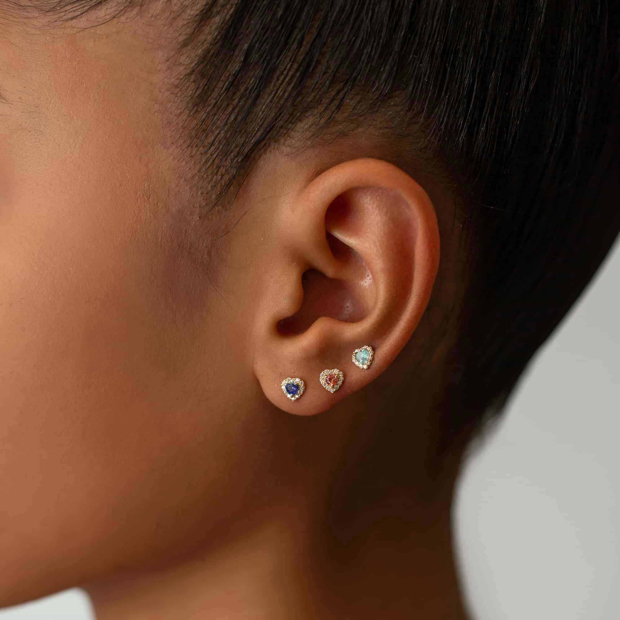 14K gold heart-cut aquamarine halo stud shown on a model's ear among blue, peach and teal studs