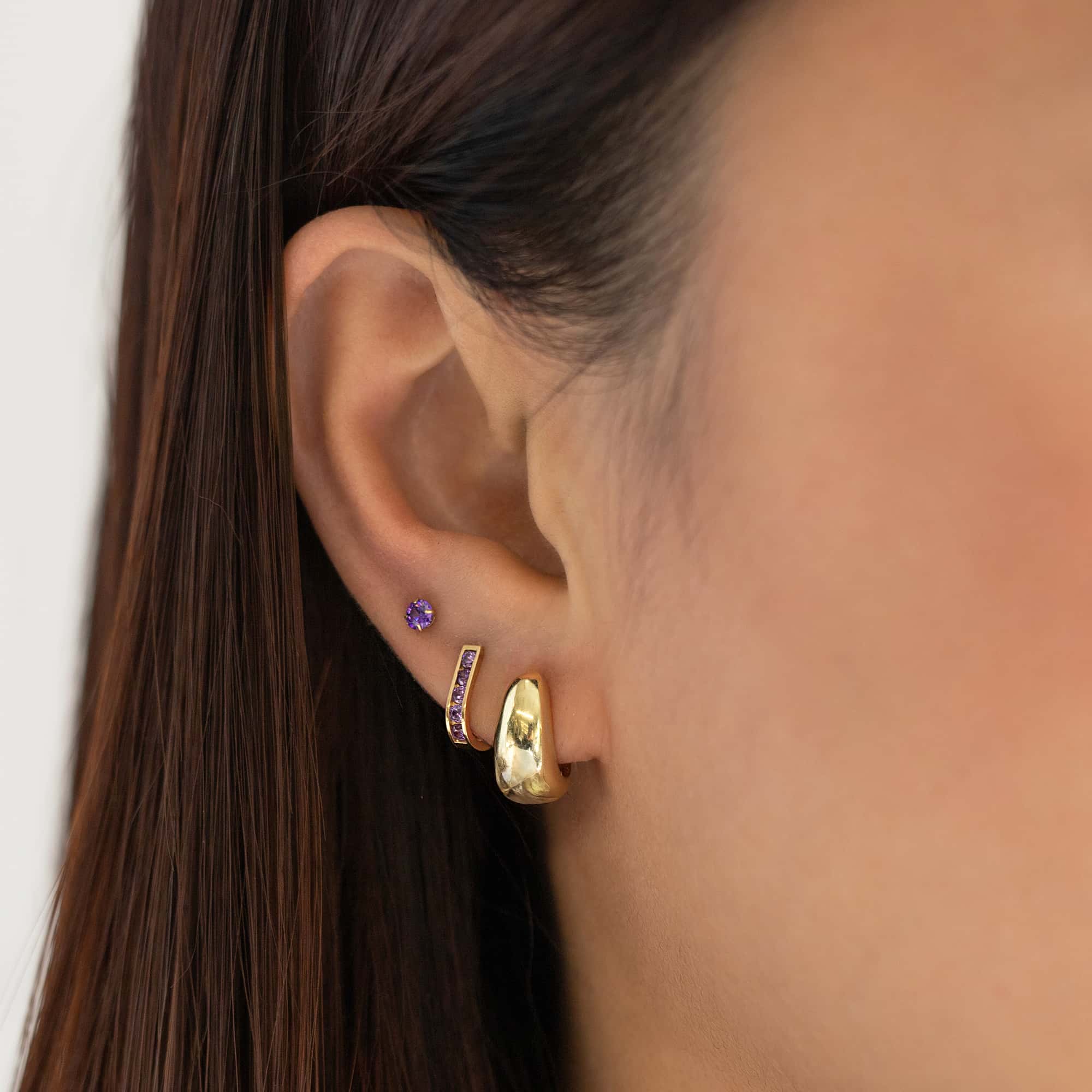 Gold purple-stone stud and a domed teardrop hoop with a purple-stone bar huggie on a model's ear