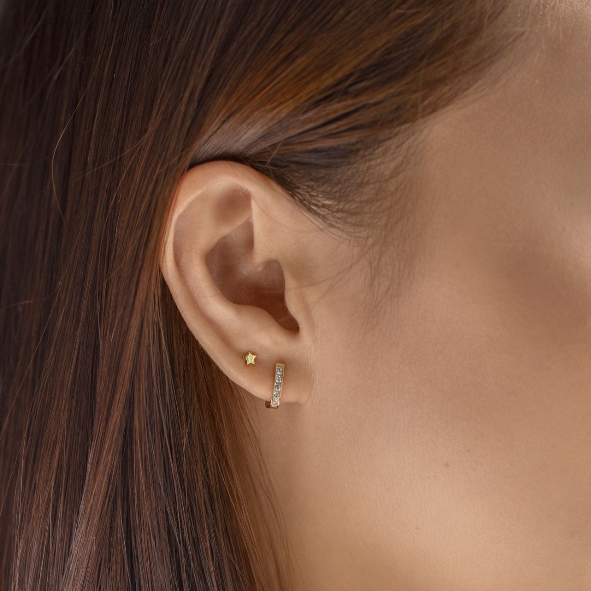 14K gold blue zircon huggie with clear stones shown on a model's lobe beside a tiny gold star stud