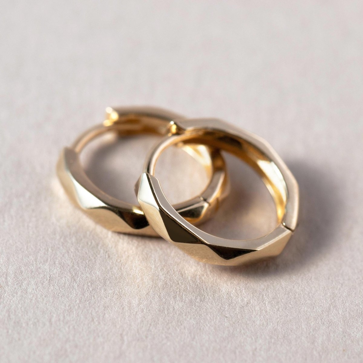 Pair of 14K gold faceted mini huggie hoops, close-up detail on a cream background