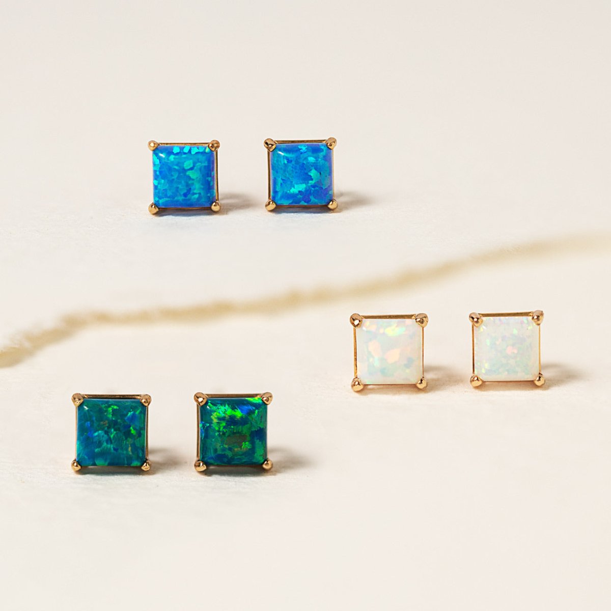 Three pairs of 14K gold square opal-look studs in blue, green and white, on cream marble