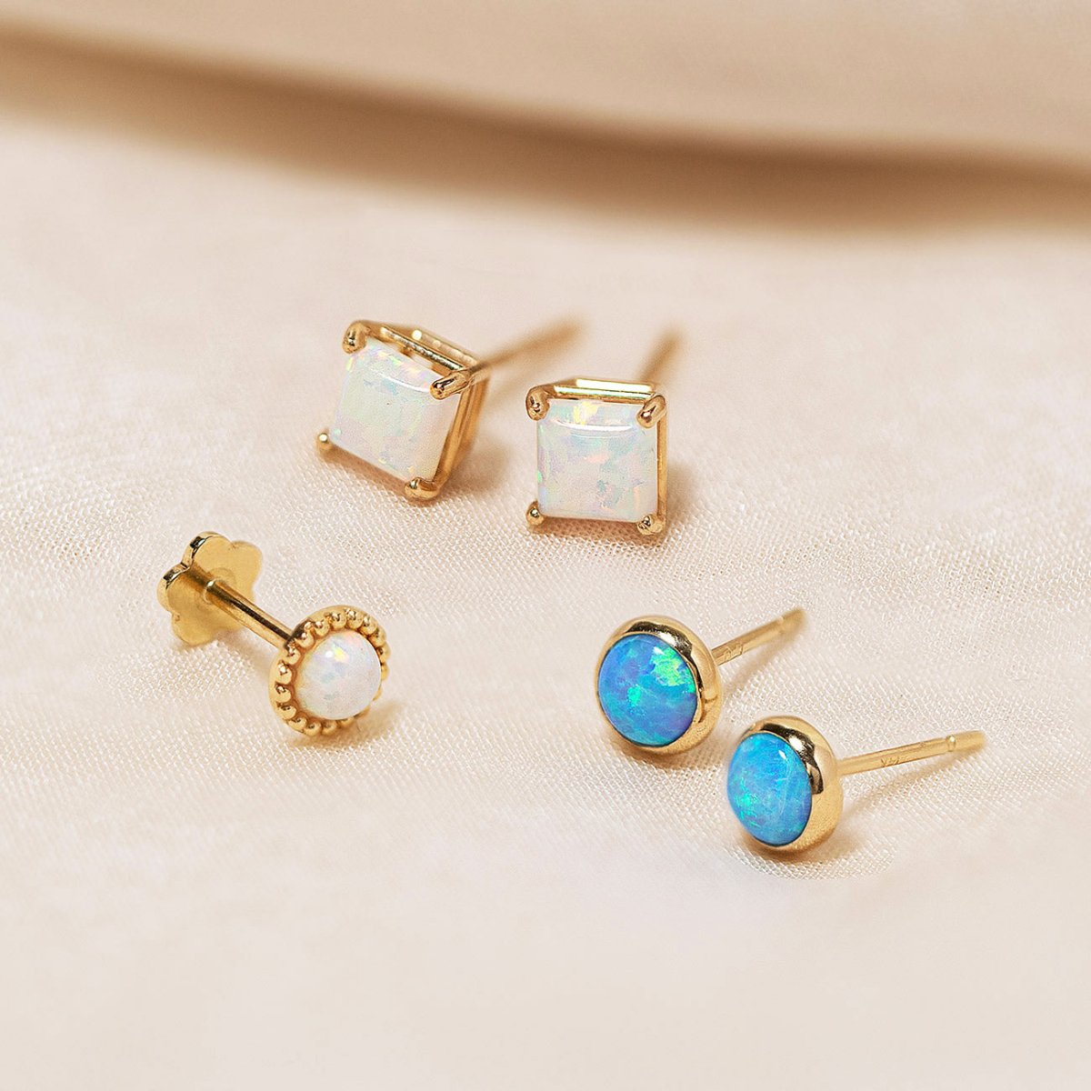 14K gold opal flat back studs in square, beaded round and round bezel styles, on a cream background