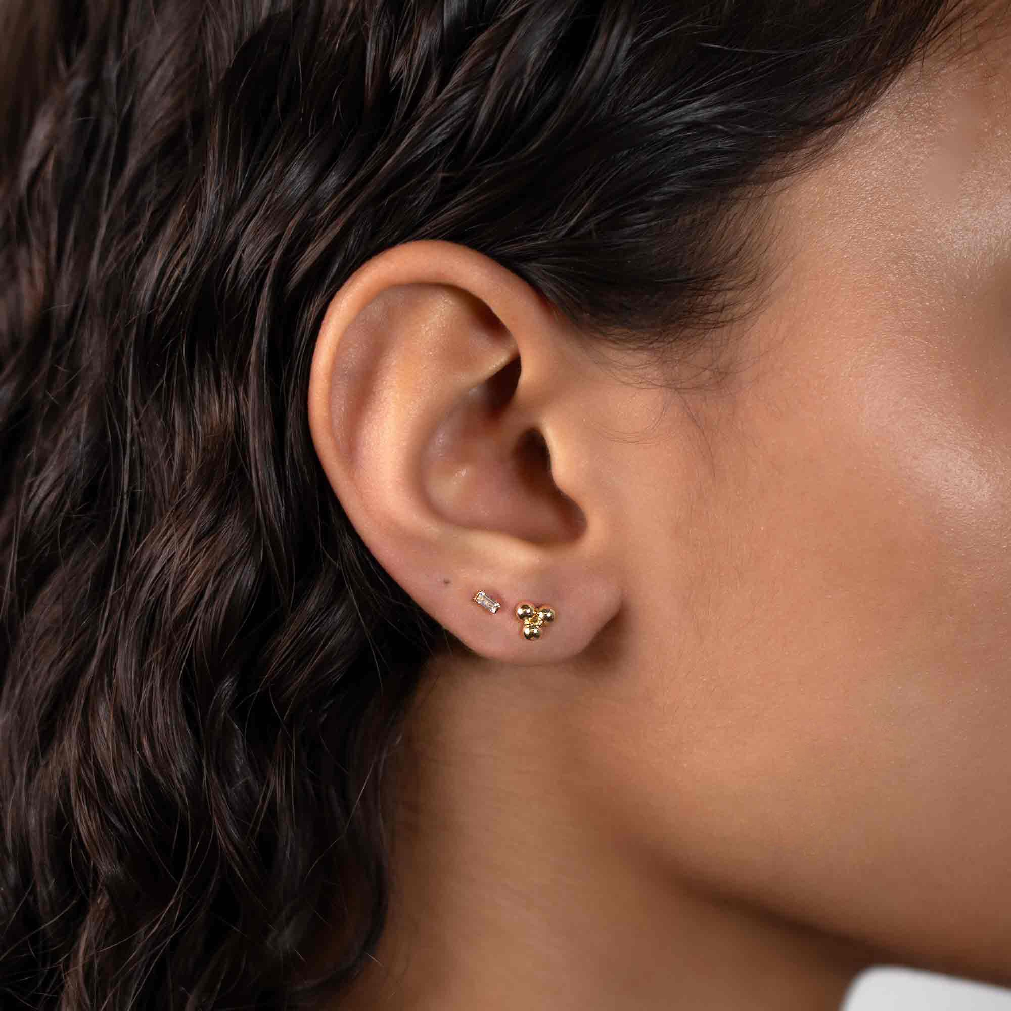 14K gold baguette stud with a clear stone, shown on a model's lobe beside a gold cluster stud
