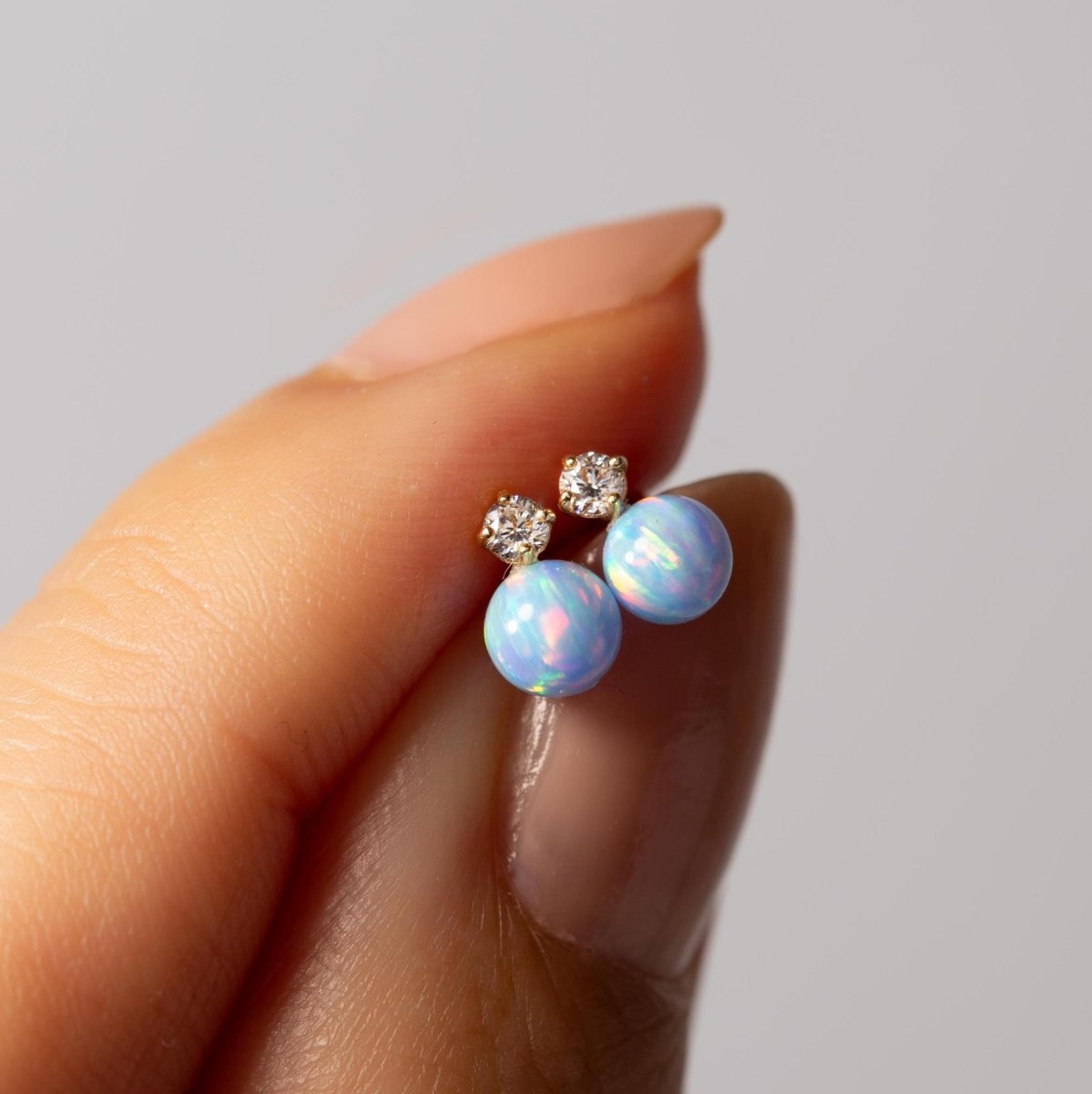 14K gold light blue opal ball studs with clear accent stones, held between fingertips for scale