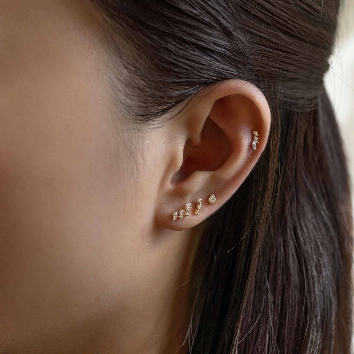 14K gold bezel pear single stud with a clear stone, shown on a model's lobe in an ear stack