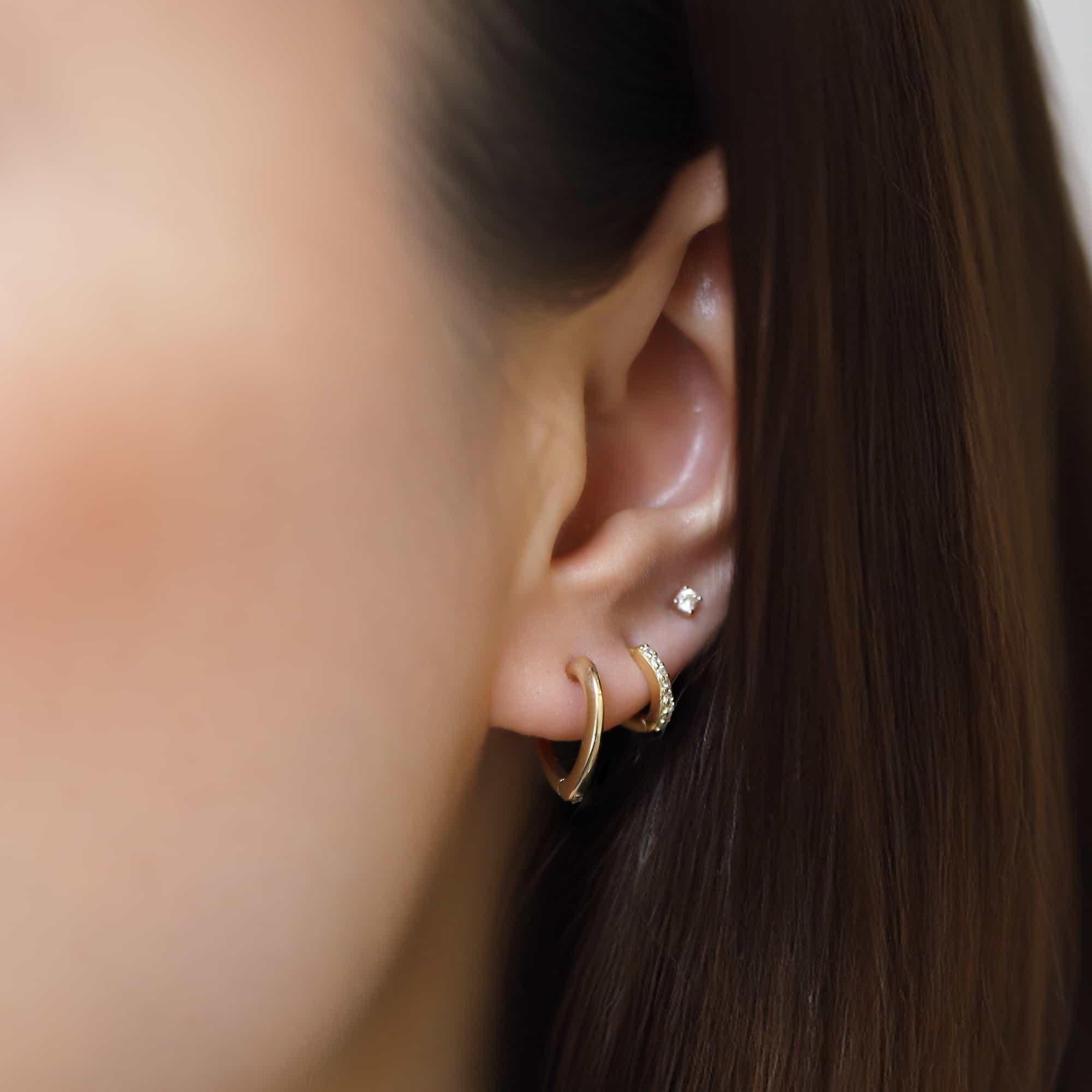 14K gold mini heart huggie hoop worn with a plain hoop and clear-stone stud on a model's lobe