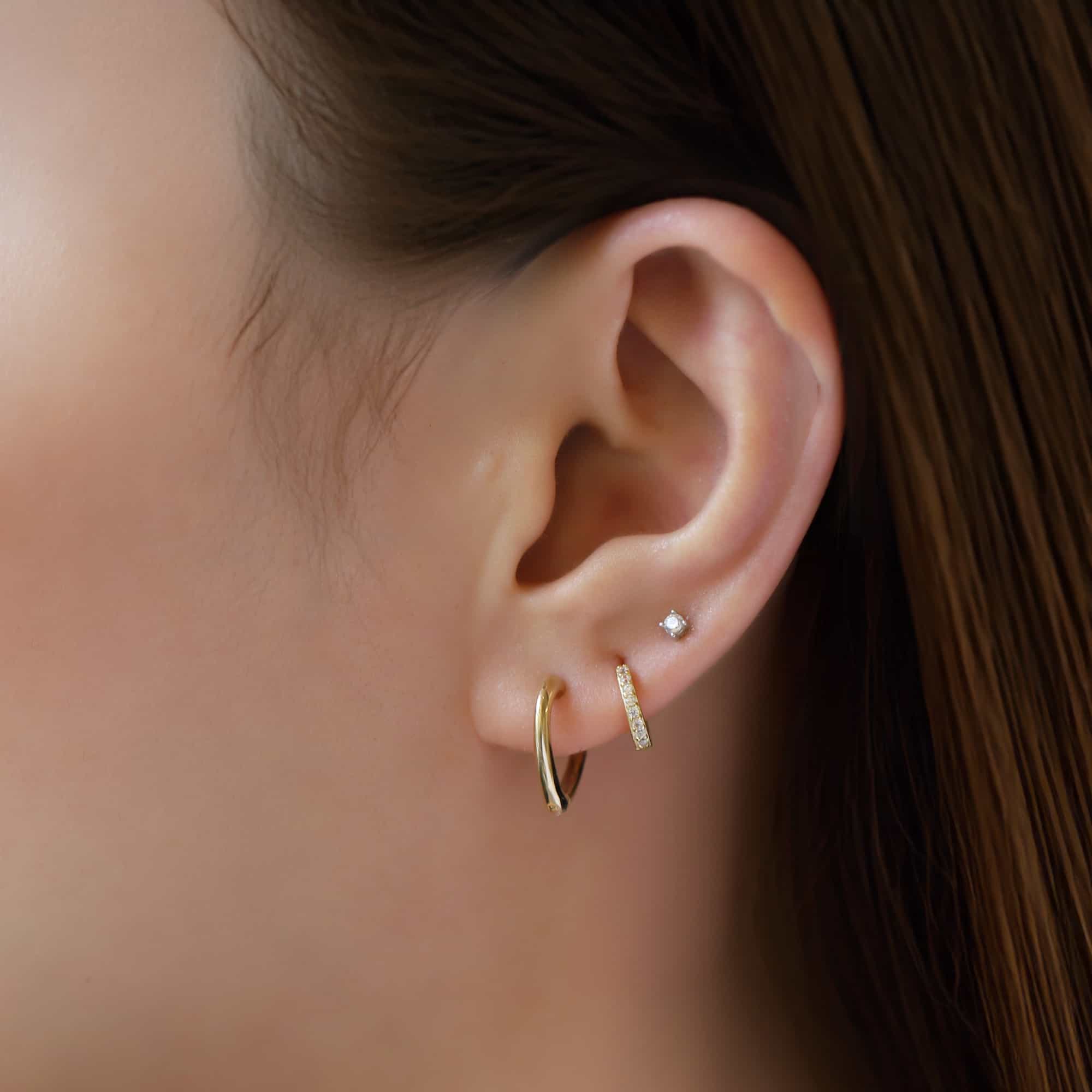 14K gold mini heart huggie hoop shown on a model's lobe in an ear stack with a tiny clear stud